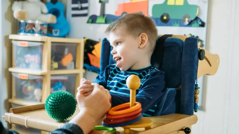 A young boy with cerebral palsy sits in a supportive chair and plays with stacking rings while an adult holds his hand